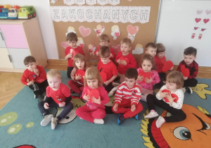 Wszystkie dzieci siedzą pod tablicą, pozują do zdjęcia przykładając do serca czerwone serduszka.