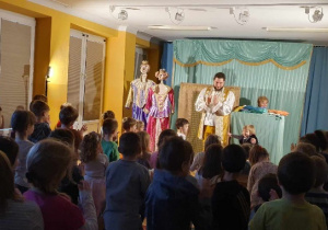 Na scenie stoją dwa maluszki wraz z aktorem, a pozostałe dzieci także stoją i klaskają.