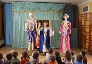 Aktorka wraz z kukłami króla i królowej stoją na środku sali.
