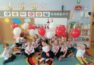 Dzieci siedzą na dywanie prezentując balony w kolorach narodowych z okazji Święta Niepodległości.