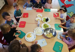 Dzieci siedzą w kole przy stolikach i wspólnie przygotowują sałatkę owocową z okazji Dnia Zdrowego Jedzenia i Gotowania.