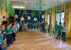 Dzieci z całego przedszkola stoją w półkolu na sali gimnastycznej i śpiewają piosenkę dla Pani Wiosny.