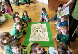 Dzieci z II grupy siedzą wokół dużego zielonego bristolu, prezentując ułożony z kartek A4 wzór Pani Wiosny.