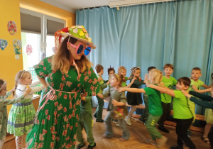 Pani Wiosna w okularach, kapeluszu i zielonej sukience w kwiaty, prowadzi dzieci w pociągu.