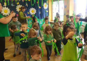 Dzieci stoją na sali gimnastycznej, w ręku trzymają paski zielonej bibuły i poruszają nimi w różnych kierunkach.