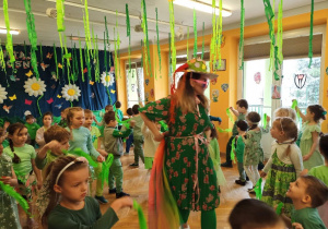 Pani Wiosna ubrana w okulary, kwiecisty kapelusz i zieloną sukienkę w kwiaty, przechadza się wokół przedszkolaków tańczących z zieloną bibułą w ręku.