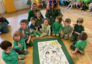 Dzieci z I grupy wraz z Paniami siedzą wokół dużego zielonego bristolu, prezentując ułożony z kartek A4 wzór Pani Wiosny.