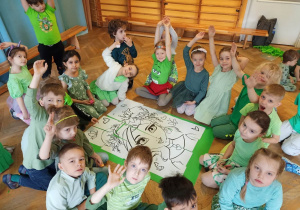 Dzieci z IV grupy siedzą wokół dużego zielonego bristolu, prezentując ułożony z kartek A4 wzór Pani Wiosny.