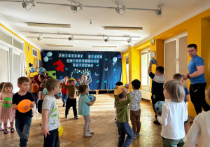 Dzieci spacerują po sali z piłkami lub krążkami gimnastycznymi w ręku.