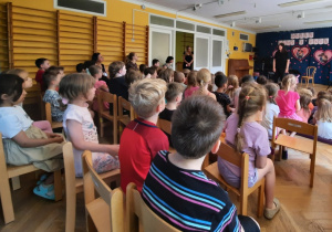 Dzieci siedzą na sali gimnastycznej i słuchają wprowadzenia do tematyki koncertu.