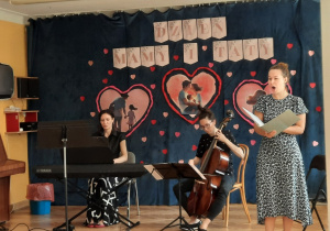 Na środku sali wystepują śpiewaczka operowa, klawesynistka oraz artystka grająca na viola da gambie.