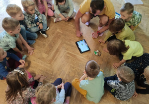 Dzieci siedzą w kole między nimi instruktor robotyki. Po środku leży tablet. Dzieci uważnie słuchają na czym polega programowanie.