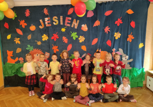 Dzieci stoją pod jesienna dekoracja z napisem Jesień. Ubrane w kolory jesieni uśmiechają się do zdjęcia.