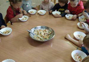 Dzieci siedzą przy stole. Na środku miska z sałatką, a przed nimi pełne salaterki sałatki. Wspólnie jedzą gotową potrawę.