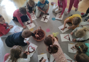 Dzieci siedzą na dywanie, na środku kosz z kasztanami dzieci układają z kasztanów wzory wg instrukcji.