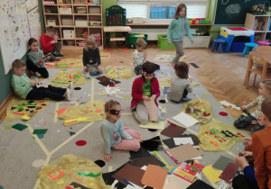 Dzieci siedzą na dywanie, przed nimi porozkładane worki foliowe. Ozdabiają kolorowymi wycinkami "deszczową pelerynę".