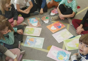 Dzieci siedzą w kole na dywanie demonstrując swoje rysunki.