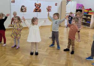 Dzieci uczestniczą w rozgrzewce - stoją w rozsypce z rękami podniesionymi ku górze.