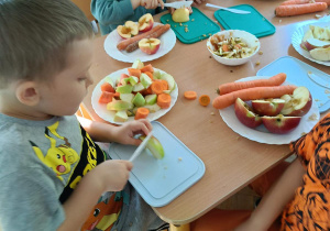 Trzech chłopców siedzi przy stoliku i kroi jabłka oraz marchewki na kawałki.