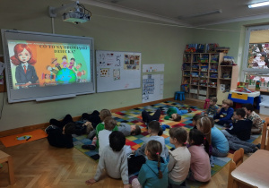 Dzieci siedzą na dywanie i oglądają prezentację z okazji Międzynarodowego Dnia Praw Dziecka.