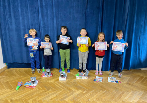 Sześcioro dzieci z P9 stoi na sali gimnastycznej prezentując dyplomy, a przed nimi położone są nagrody i upominki za udział w konkursie.