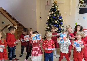 Maluchy stoją przy choince w holu przedszkola, trzymając w rączkach upominki pozostawione tam przez Mikołaja.