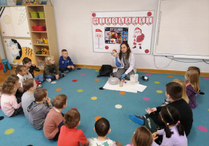 Dzieci siedzą na dywanie w półkolu i witają się z prowadzącą warsztaty chemiczne LABOLO i kukiełką profesorkiem Labolkiem.