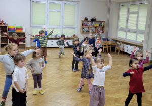 Maluszki stoją w rozsypce na sali. Podnoszą ręce do góry i poruszają nimi na boki.