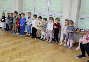 Dzieci stoją w jednym rzędzie i słuchają z uwagą instrukcji prowadzącej.