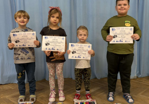 Dziewczynka i trzech chłopców stoją w rzędzie i pozują do grupowego zdjęcia z nagrodami i dyplomami za udział w konkursie.