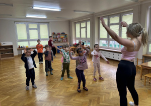 Dzieci stoją w rozsypce na sali i wymachują rękami uniesionymi do góry podczas zajęć zumby.