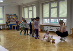 Dzieci stoją ustawione w pociąg na sali, a dziewczynka przechodzi pod kijem, bawiąc się w "Limbo".