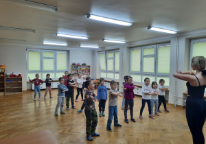 Dzieci stoją w rozsypce na sali podczas zajęć zumby i powtarzają kroki taneczne za prowadzącą zajęcia.