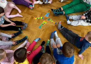 Dzieci wspólnie z nauczycielką siedzą w kole na podłodze prezentując swoje kolorowe skarpetki nie do pary.