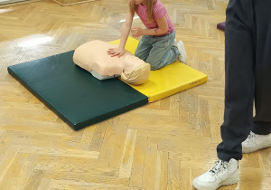 Dziewczynka klęczy na materacu z jedną ręką na manekinie i ćwiczy prawidłowy masaż serca.