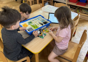 Dziewczynka i dwóch chłopców siedzi przy stoliku i budują konstrukcję z klocków Lego na warsztatach z robotyki.