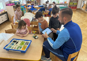 Dzieci siedzą przy stoliku i z pomocą prowadzącego programują zbudowany pojazd z klocków Lego za pomocą programu na tablecie.