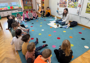 Dzieci siedzą na dywanie w półkolu, a prowadząca warsztaty pokazuje im probówkę z drobinkami złota, które powstały podczas eksperymentu.