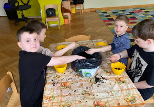 Czworo chłopców siedzi przy stoliku i przekładają ziemię do doniczek w kształcie skorupki jajka.