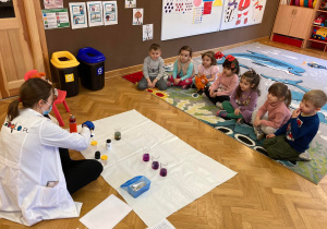 Pani instruktor pokazuje dzieciom siedzącym na dywanie eksperyment chemiczny z płynami.