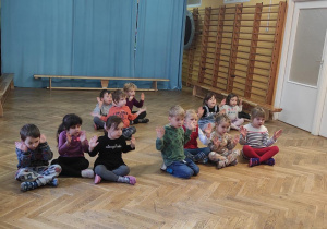 Dzieci siedzą po turecku na podłodze i słuchają uważnie pani instruktor tańca.