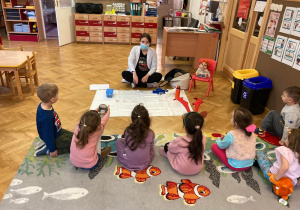 Pani instruktor tłumaczy dzieciom o czym będą zajęcia chemiczne. Dzieci siedzą na dywanie w rzędzie.