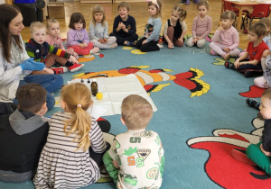 Dzieci siedzą na dywanie w półkolu i rozmawiają z prowadzącą warsztaty o przeprowadzonym doświadczeniu.