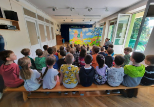 Przedszkolaki siedzą na ławeczkach, a przed nimi stoi dekoracja przedstawiająca fiołkową krainę.