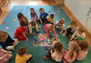 Przedszkolaki siedzą wokół plakatu, jedna z dziewczynek przykleja Śmieszkę na Wyspie Radości.