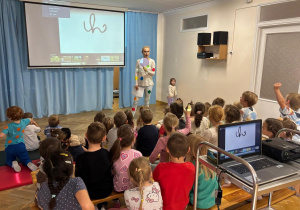 Dzieci siedzą zwrócone twarzą do ekranu, przed nimi stoi Pani nauczycielka i wprowadza dzieci w tematykę dnia.