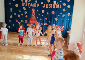 Maluchy stoją w półkolu trzymając się za ręce, w tle dekoracja z okazji Powitania Jesieni.