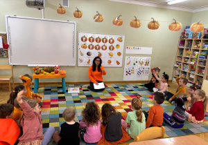 Dzieci siedzą na dywanie w półkolu i rozmawiają z nauczycielką nt. dyni, np. do czego jej używamy.
