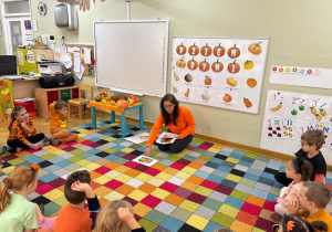 Dzieci siedzą na dywanie w półkolu, a nauczycielka prezentuje im fotografię potraw, które można wykonać z dyni.