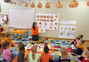 Dzieci siedzą na dywanie w półkolu, a nauczycielka prezentuje im budowę dyni.